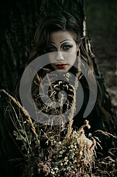 Portrait of a young woman in the Gothic gloomy image of a witch in the forest with a bouquet. Halloween costume