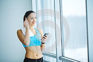 Portrait of young woman in gym posing with towel on neck and listening to music using earphones and smartphone,