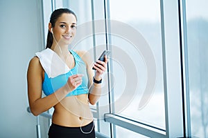 Portrait of young woman in gym posing with towel on neck and listening to music using earphones and smartphone,