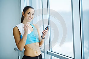 Portrait of young woman in gym posing with towel on neck and listening to music using earphones and smartphone,