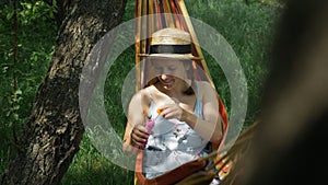 Portrait of young woman in hat blowing bubbles in hammock at green garden. Happy smiling girl in dress resting in hammock and blow
