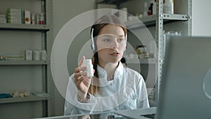 Portrait of young woman in headset, showing pills in jar to client, in pharmacy