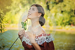 Portrait of a young woman in a historical dress from the 19th century. Beautiful girl with a rose in her hand. Summer background.