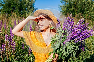 Portrait of young woman holding bouquet of lupin flowers walking in summer meadow. Stylish girl picking blooms