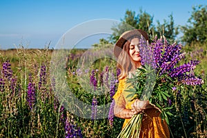 Portrait of young woman holding bouquet of lupin flowers walking in summer meadow. Stylish girl picking blooms