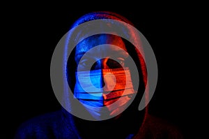 Portrait of a young woman in a hood, wearing a protective medical mask, illuminated by red and blue light, isolated on black backg