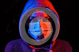Portrait of a young woman in a hood, wearing a protective medical mask, illuminated by red and blue light, isolated on black backg