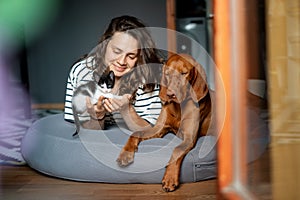 Portrait of a young woman with a Hungarian Pointer dog and a small kitten in her arms lying at home in a room on a bag