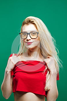 Portrait of Young Woman with Long Blond Hair, Eyeglasses and Bare Shoulders in Red Top Posing on Green Background in