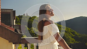 Portrait of young woman in a long dress, standing on the villa& x27;s terrace or balcony looking on a mountain sunset