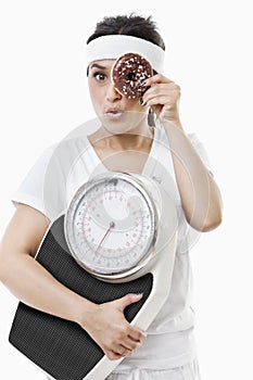 Portrait of young woman looking through donut holding weight scale over white background