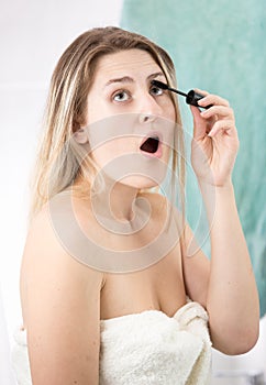 Portrait of young woman opens mouth while applying mascara and doing makeup in bathroom