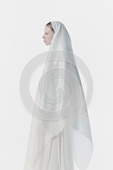 Portrait of young woman with pale skin and blonde hair, wrapped in white fabric looks as nun against white studio