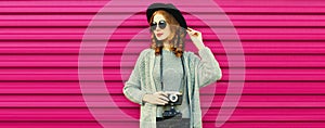 Portrait of young woman photographer with film camera wearing knitted sweater cardigan, black round hat on red background