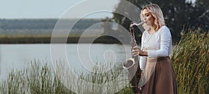 Portrait of young woman playing saxophone on bank of the river in reeds, girl with woodwind musical instrument on nature