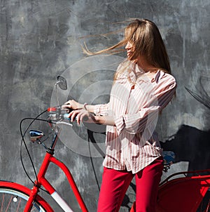 Portrait young woman in red chino holding hands on her red vintage bicycle, standing against gray wall. The wind blows her hair. O