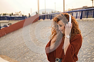 Portrait of young woman, red hair, freckles, with red sweater and white headphones, listening to music, in an outdoor park .