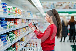 Portrait of young woman scans the QR code on a package of yogurt. In the background, a supermarket with visitors in a