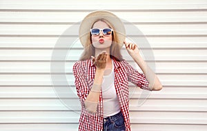 Portrait young woman sending sweet air kiss blowing red lips in summer round straw hat, checkered shirt on white wall