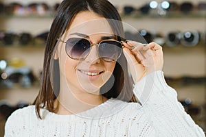 Portrait of a young woman shopping, standing in store and trying sunglasses near a mirror