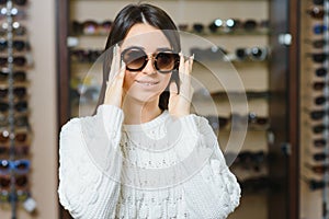 Portrait of a young woman shopping, standing in store and trying sunglasses near a mirror