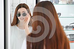 Portrait of a young woman shopping, standing in store and trying sunglasses near a mirror.
