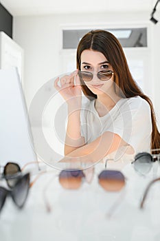 Portrait of a young woman shopping, standing in store and trying sunglasses near a mirror.