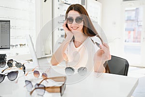 Portrait of a young woman shopping, standing in store and trying sunglasses near a mirror.