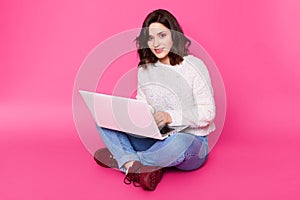 Portrait of young woman sits with legs crossed, uses laptop computer. Brunette female works online at home, earns money in