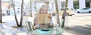 Portrait of young woman sitting in outdoor cafe, hiding behind journal, holding notebook in hands and smiling