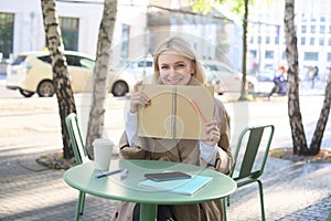 Portrait of young woman sitting in outdoor cafe, hiding behind journal, holding notebook in hands and smiling