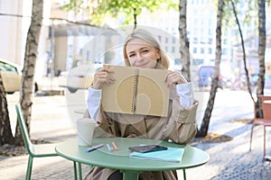 Portrait of young woman sitting in outdoor cafe, hiding behind journal, holding notebook in hands and smiling