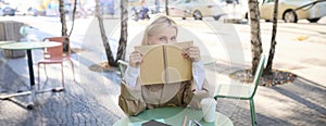 Portrait of young woman sitting in outdoor cafe, hiding behind journal, holding notebook in hands and smiling