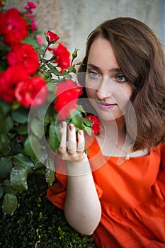 Portrait of young woman in the spring time. Red roses flowers blossoms. Girl dressed in red dress smelling roses in garden.