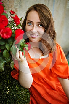 Portrait of young woman in the spring time. Red roses flowers blossoms. Girl dressed in red dress smelling roses in garden.