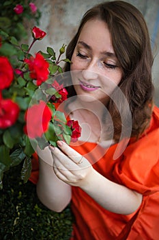 Portrait of young woman in the spring time. Red roses flowers blossoms. Girl dressed in red dress smelling roses in garden.