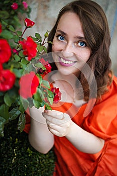 Portrait of young woman in the spring time. Red roses flowers blossoms. Girl dressed in red dress smelling roses in garden.