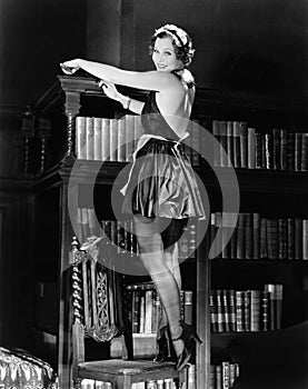 Portrait of a young woman standing on a chair and dusting a bookshelf in a outfit