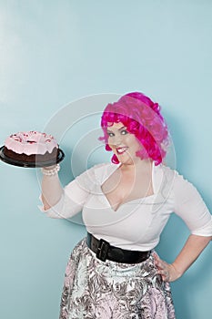 Portrait of young woman standing with hands on hips holding cake over colored background