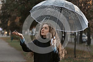 Portrait of young woman standing under transparent umbrella to see if it is raining. Girl with brown hair walks in park