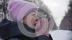 Portrait of a young woman on the street who breathes in her arms to keep warm while walking in a winter park. Slow