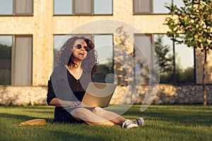 Portrait of young woman in sunglasses sitting on green grass and laugh, using laptop.