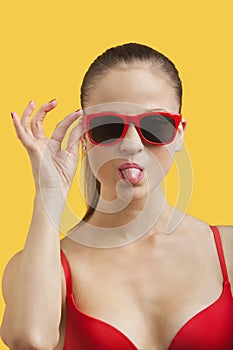 Portrait of young woman in sunglasses sticking out tongue over yellow background