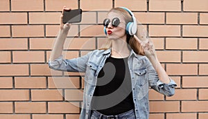 Portrait young woman taking selfie picture by smartphone in wireless headphones listening to music wearing a black round hat