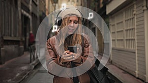 Portrait of young woman teenager with headphones listening to music on phone, sing and funny dancing in street on old