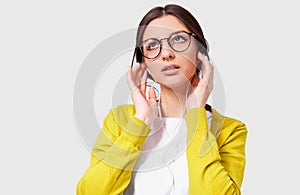 Portrait of young woman in transparent eyeglasses wearing casual outfit listening to the music from earphones. Studio shot of