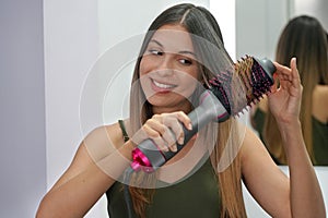 Portrait of young woman using round brush hair dryer to style hair in an easy way at home. Girl with electric blowout brush hair