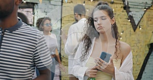 Portrait of young woman using smartphone chatting online standing at outdoor party in open air cafe