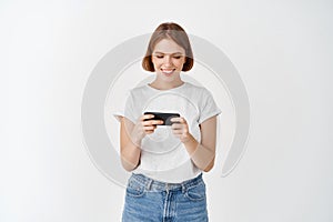 Portrait of young woman watching video on smartphone, holding phone horizontally to play video game, white background