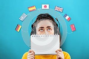 Portrait of a young woman wearing headphones, holding a notebook and looking up at the flags of different countries